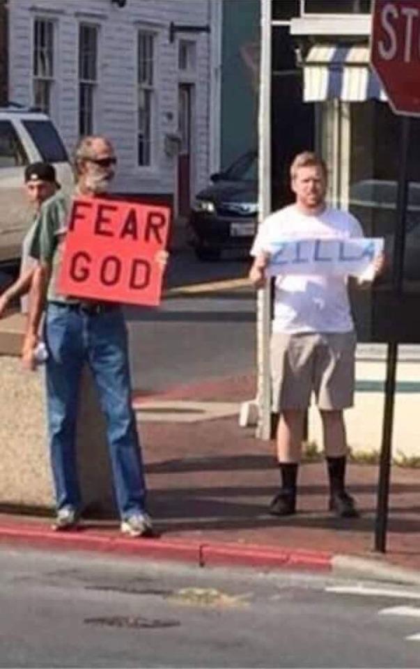 One man holds a sign saying fear god. The man standing next to him holds a sign that says zilla
